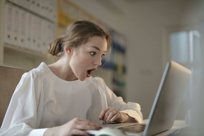 Young woman shocked while looking at laptop screen, reacting to worst celebrity wax figures ever made online.