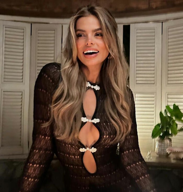 Brooks Nader smiling indoors wearing a black patterned top with cut-out details and long wavy hair styled loosely.