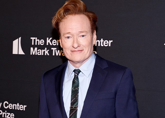 Conan O’Brien wearing a navy suit and patterned tie at a media event with a dark backdrop and white text.