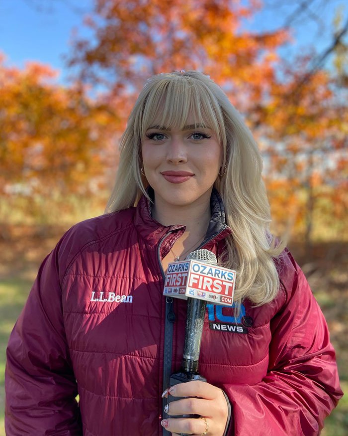 Curvy news anchor outdoors holding Ozarks First microphone with fall trees in the background during a broadcast.