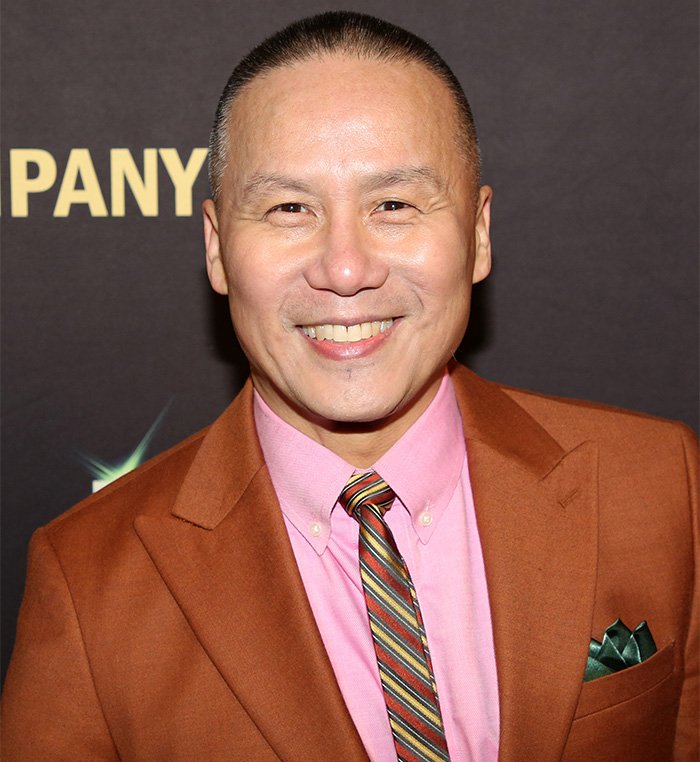 BD Wong, Jurassic World star, smiling in a brown suit and striped tie at a public event against a dark backdrop.