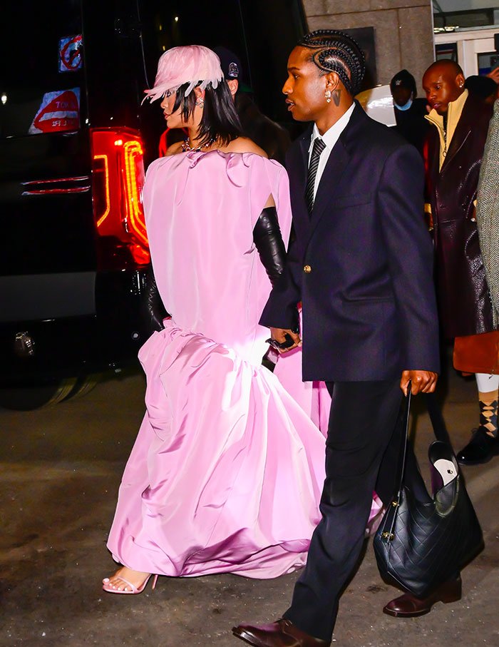 Rihanna in a flowing pink dress and hat, accompanied by a man carrying a large black bag at a red carpet event.