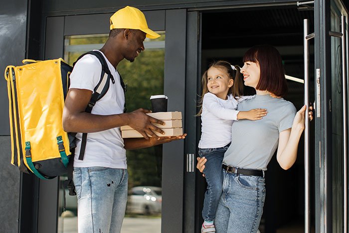 Babysitter holding pizza boxes while greeting a mother and child at the doorway during a delivery.
