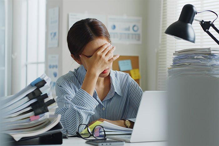Woman stressed at office desk cluttered with paperwork, upset by blasting Christmas music in the workplace environment.