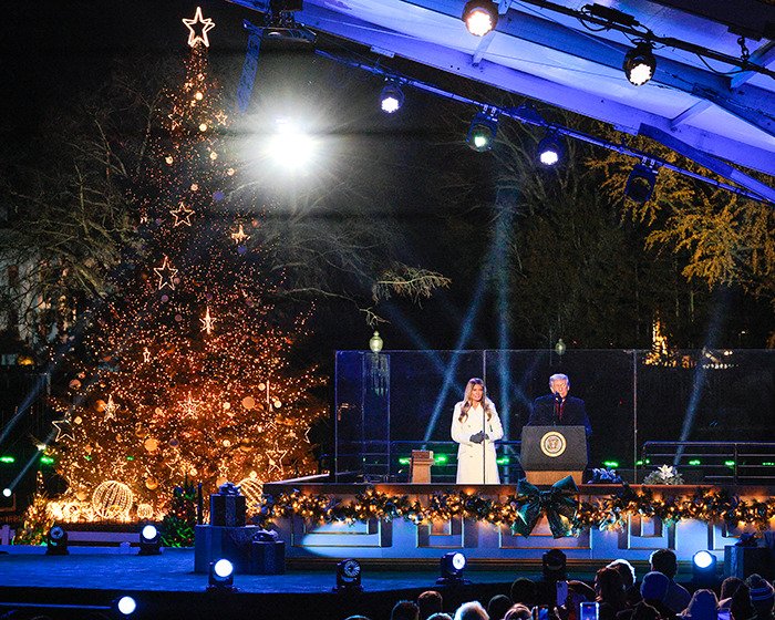 Melania standing on stage at a festive tree lighting event with bright lights and Christmas decorations around.