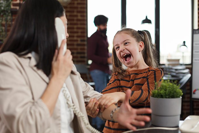 Mom struggling to handle 7-year-old child at work, showing tense interaction during phone call in office setting.