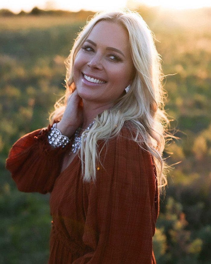 Blonde Netflix star and mom of three smiling outdoors at sunset, wearing a rust-colored dress and jewelry.