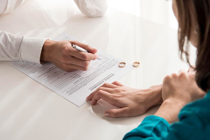 SAHM looking distressed as husband signs divorce agreement with wedding rings on the table between them.