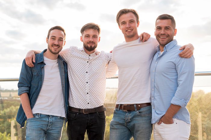 Four young men smiling and posing together outdoors, illustrating women ruining MIL&rsquo;s surprise by pretending ignorance.