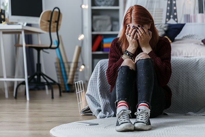 Teen girl sitting on floor in bedroom, upset and covering her face, depicting insecurity about stepmother dynamics.