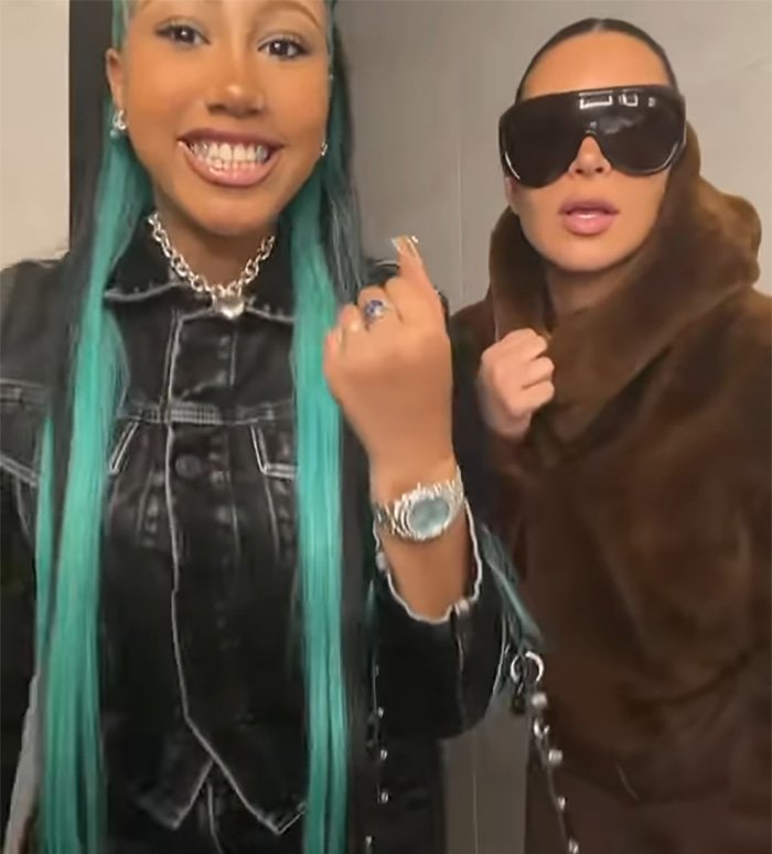 Two women pose indoors, one with turquoise hair and jewelry, the other wearing oversized sunglasses and a brown fur coat.