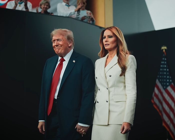 Donald and Melania Trump standing together holding hands with an American flag visible in the background.