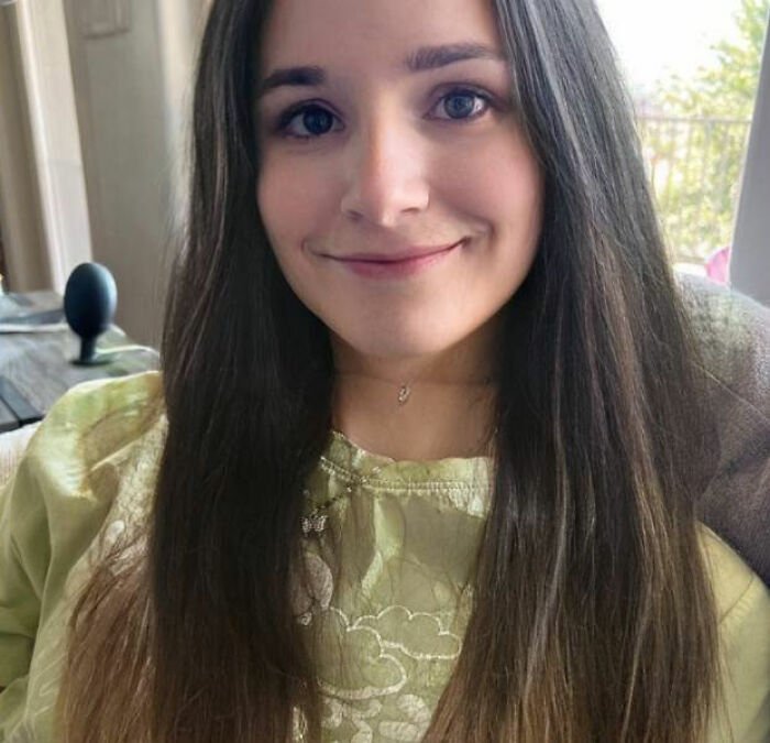 Trans Muslim woman with long dark hair and a green top smiling indoors near a window with greenery outside.