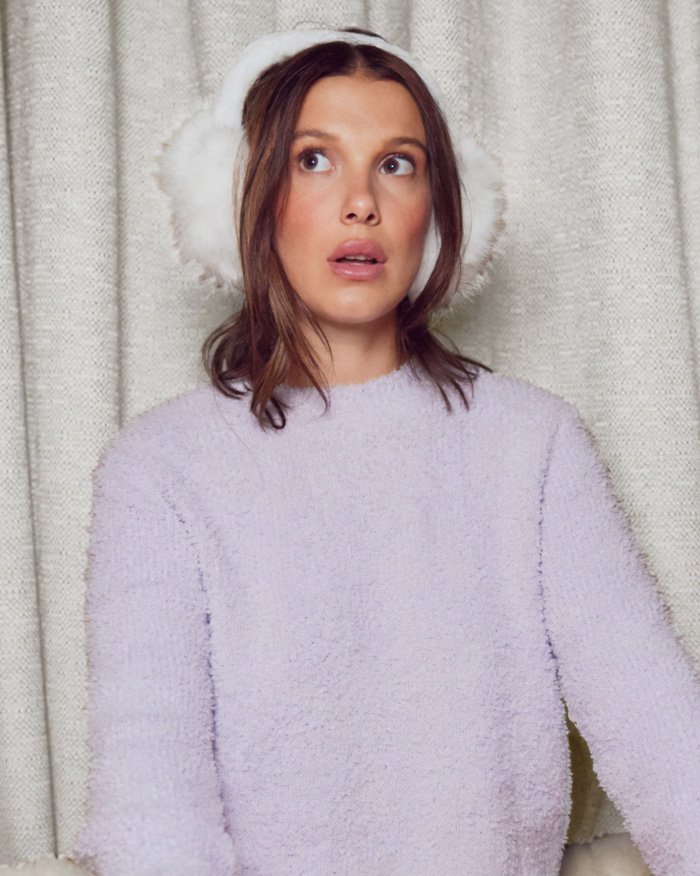 Millie Bobby Brown wearing white earmuffs and a soft lavender sweater in a casual indoor setting.