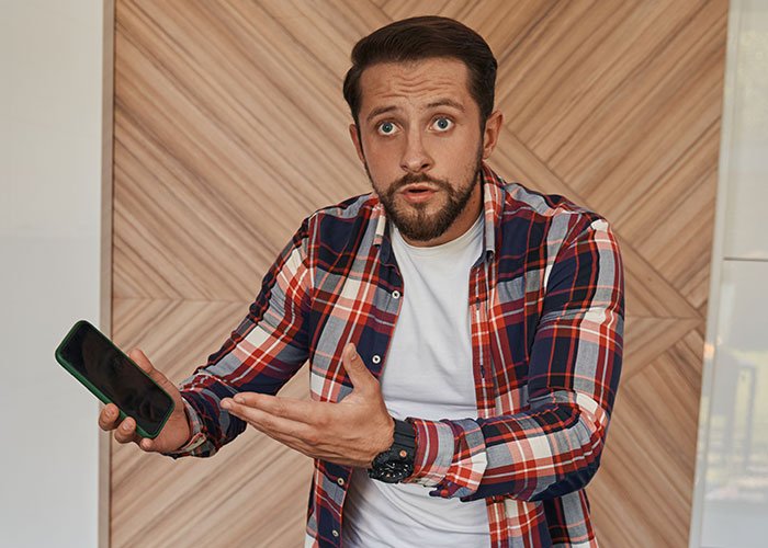 Man in plaid shirt holding phone and looking shocked, illustrating a wife ruining husband’s reputation after leaving him.
