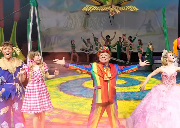 Actors in colorful costumes performing a Wizard of Oz scene on stage with vibrant lighting and a scenic backdrop.