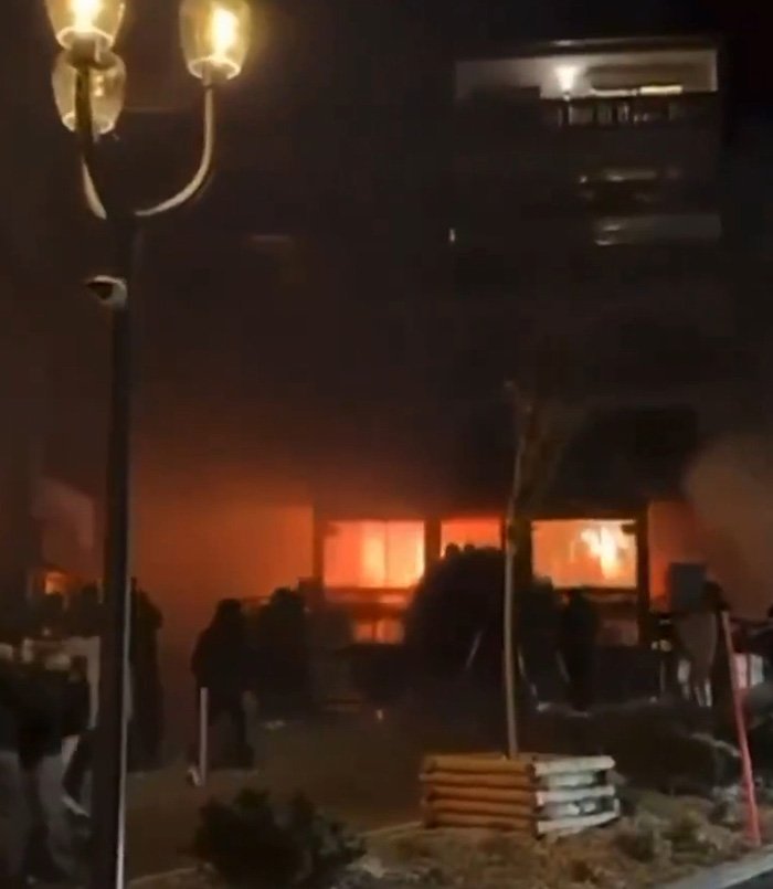 Ski resort explosion at night with bright flames visible through windows and silhouettes of people nearby.