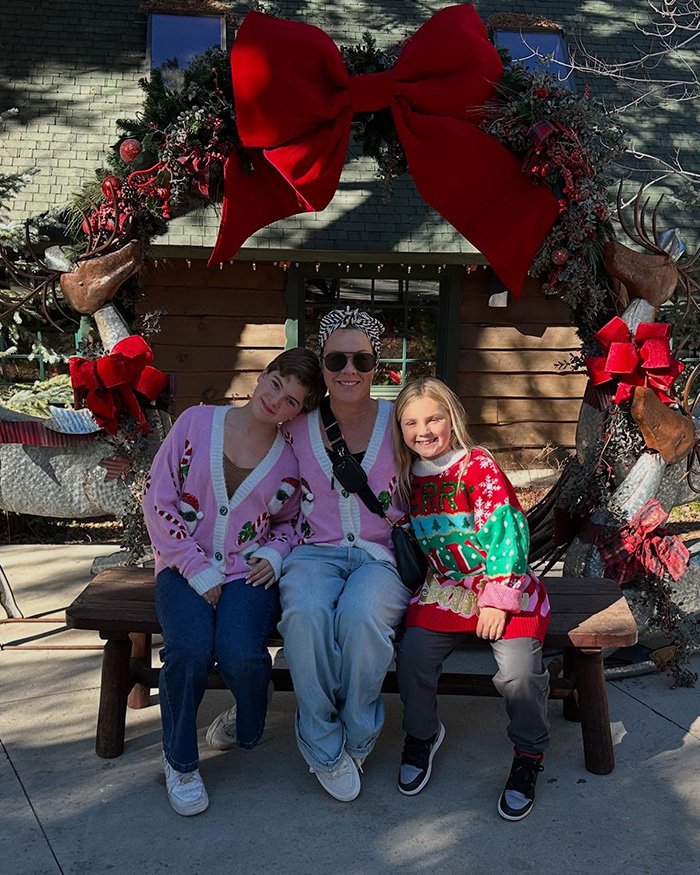 Pink with family sitting on a bench under a large festive wreath decorated with red bows and holiday ornaments.