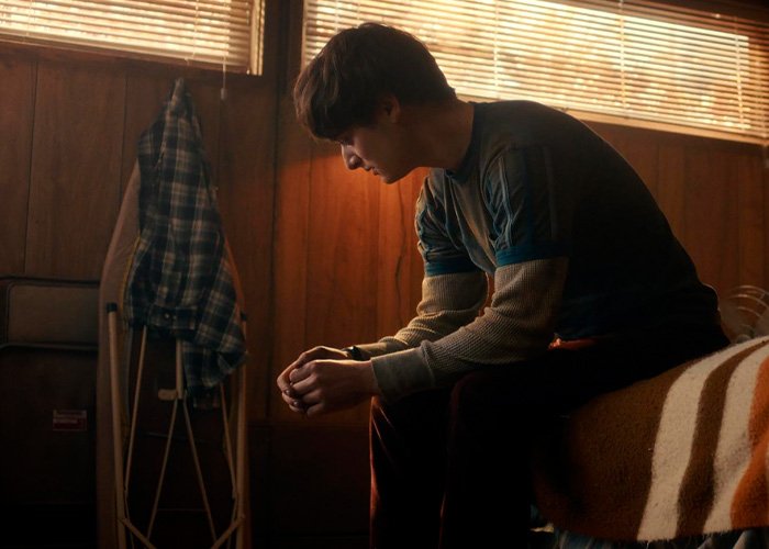 Noah Schnapp as Will Byers sitting on a bed in a dimly lit room with wooden walls and an ironing board nearby