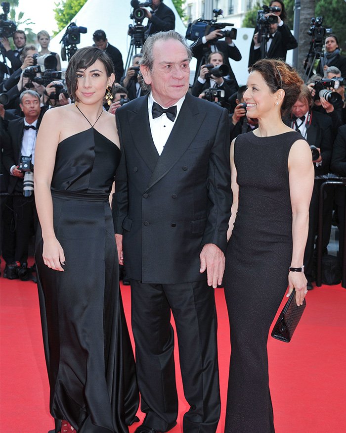 Tommy Lee Jones and two women dressed in black on a red carpet with photographers in the background.
