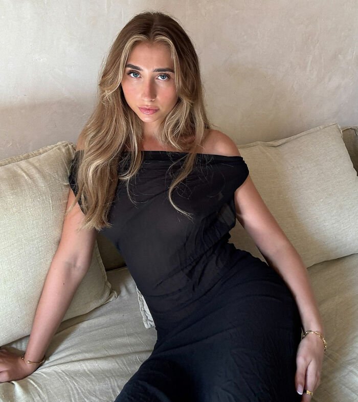 Adult star who went viral for 1000 men challenge posing on couch wearing a black dress with loose hair and natural makeup.