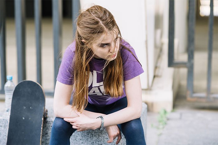 Teen girl sitting near skateboard looking distressed, illustrating fear and tension in a violent sibling relationship.