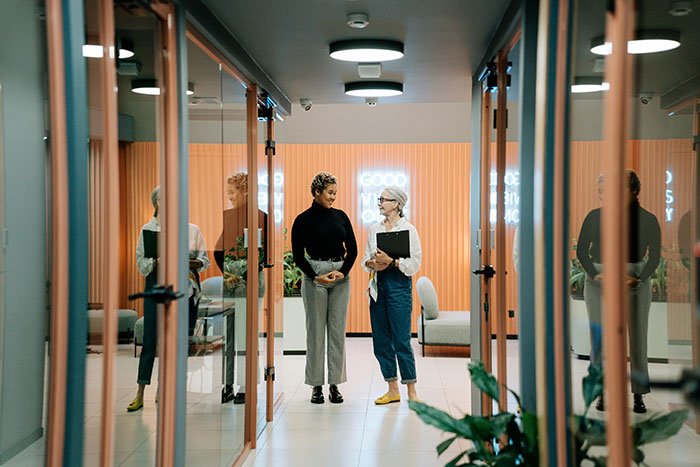 Two women coworkers having a conversation in an office hallway, illustrating a woman puts coworker on blast scenario.