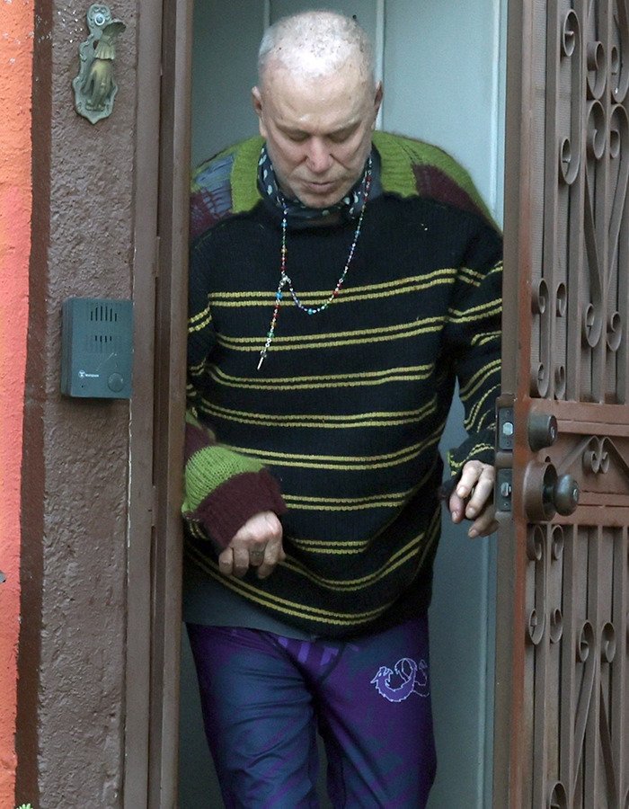Mickey Rourke wearing a striped sweater and colorful pants, exiting home amid LA eviction over unpaid rent.