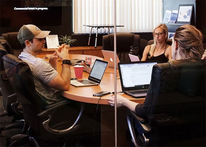 Three employees in a meeting room with laptops, discussing work hours and complaints about employee performance.
