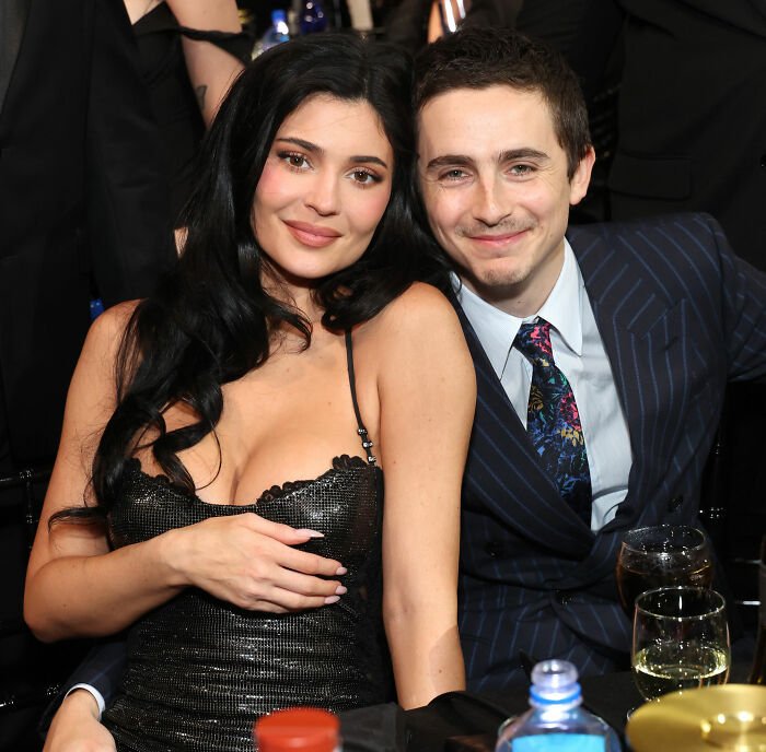 Timothée Chalamet smiling in a pinstripe suit next to a woman in a black dress at a formal event gathering.