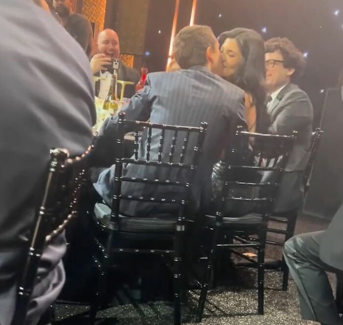 Timothée Chalamet at critics’ choice awards, sitting and standing with guests in a dimly lit event setting.