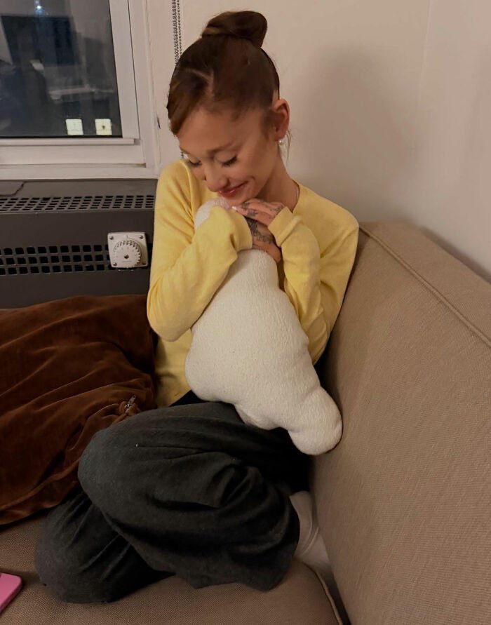 Ariana Grande sitting on a couch in casual clothes, smiling and hugging a white plush toy indoors.