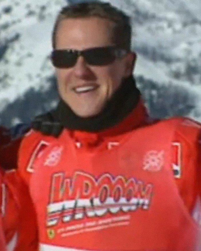 Michael Schumacher smiling outdoors in a red ski jacket and black sunglasses with snowy mountains in the background.