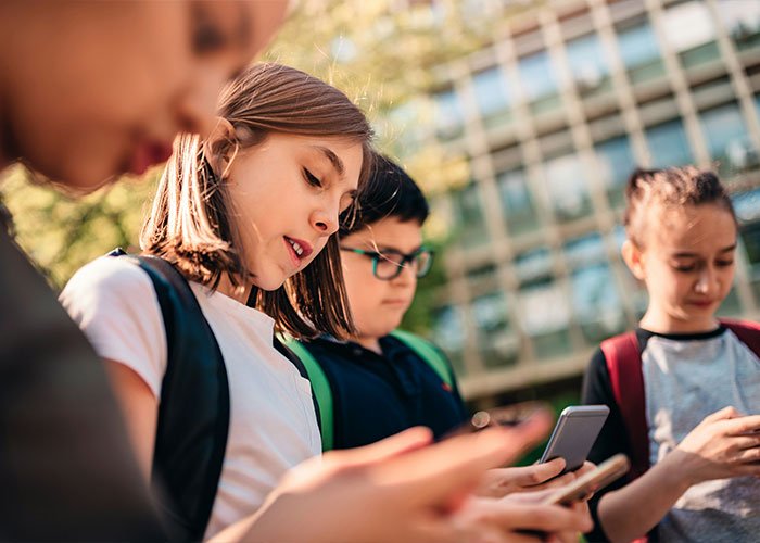 Group of young teens outdoors, focused on their smartphones, illustrating experts' views on overused slang words to banish.