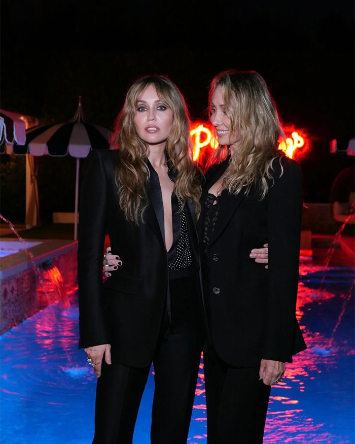 Miley Cyrus and a companion in black outfits at a nighttime red carpet event with illuminated pool background.