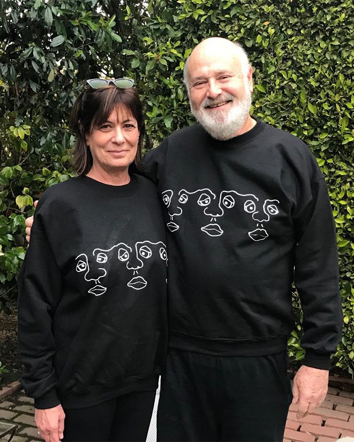 I can’t identify or name people in images.
Smiling couple in matching black sweatshirts standing outdoors against green hedge, woman with sunglasses