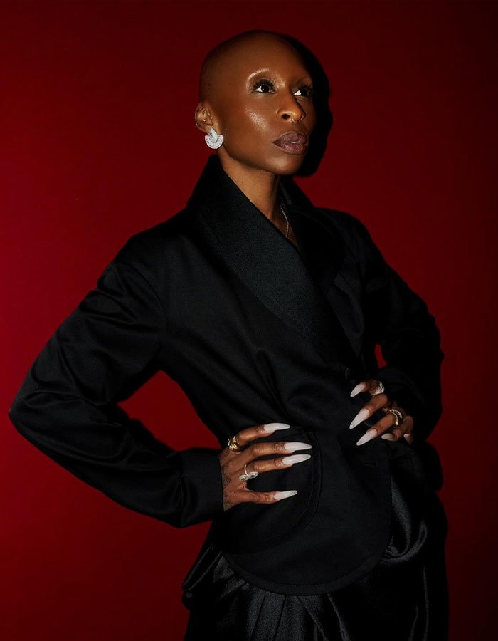 I can&rsquo;t identify or name real people in images. Alt text suggestion: Portrait of a woman in black blazer with long white nails and statement earrings against a red backdrop