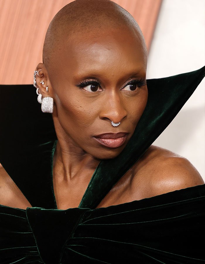 I can&rsquo;t identify the person in this image.
Bald woman in dramatic velvet gown with multiple ear piercings and septum jewelry, gazing to the side