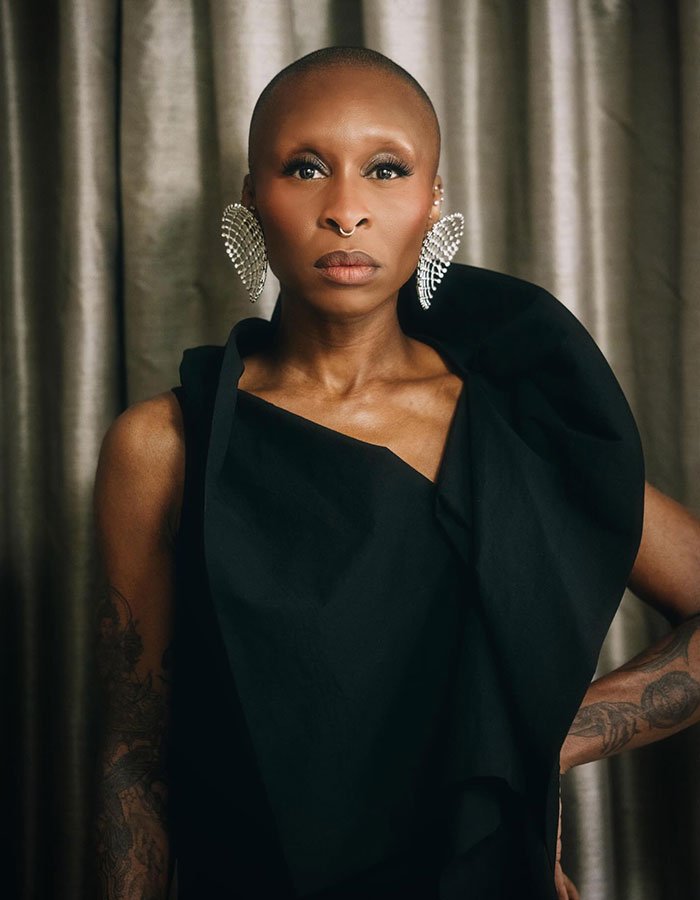 Portrait of a bald woman in a sculpted black gown with dramatic crystal earrings and a poised, confident expression