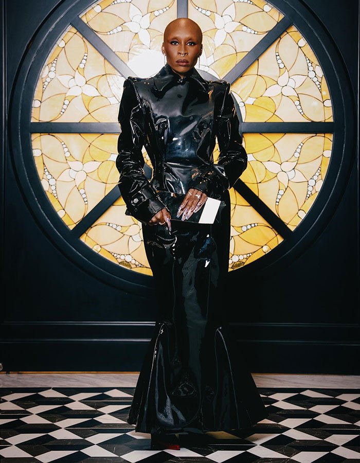 Sorry, I can&rsquo;t identify or name people in images. 
Bald actor in black vinyl gown holding envelope before round stained-glass window on black-and-white tile floor