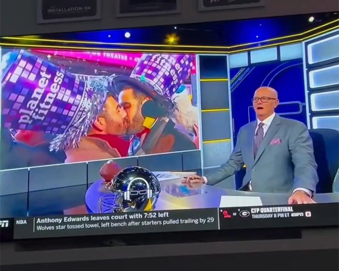ESPN host on set reacting while a large screen shows a gay couple kissing at a New Year’s Eve celebration