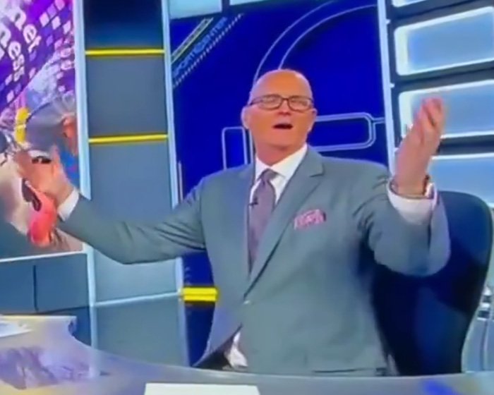 Man in TV studio wearing suit and glasses gesturing at desk, screenshot related to gay couple reaction story