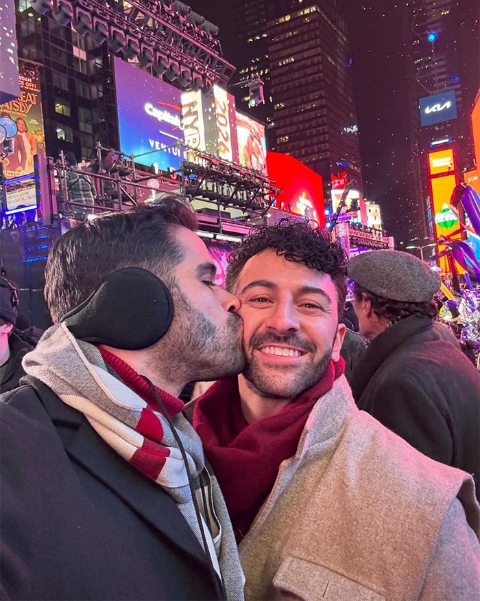 Gay couple in Times Square on New Year's Eve, one man kissing partner's cheek amid bright billboards and crowd