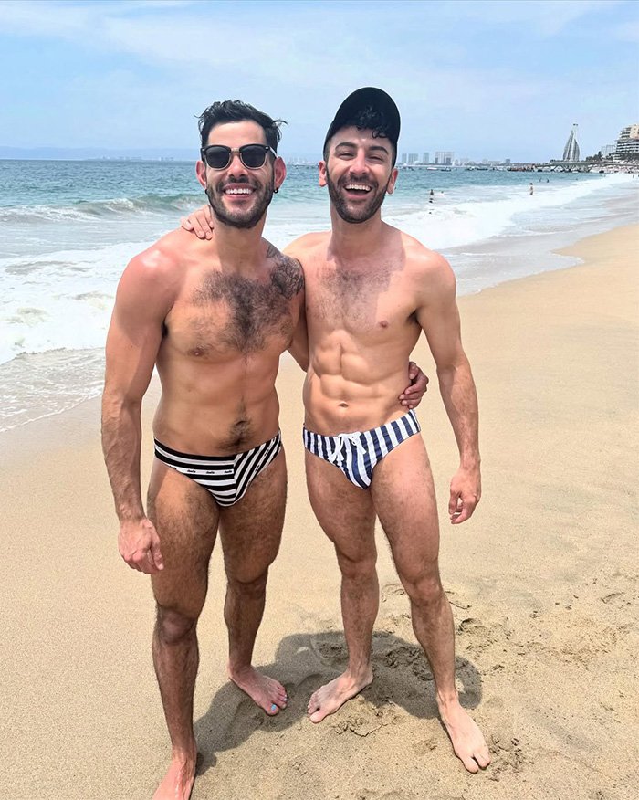 Gay couple smiling arm in arm on sandy beach wearing striped swim briefs, ocean and skyline in background