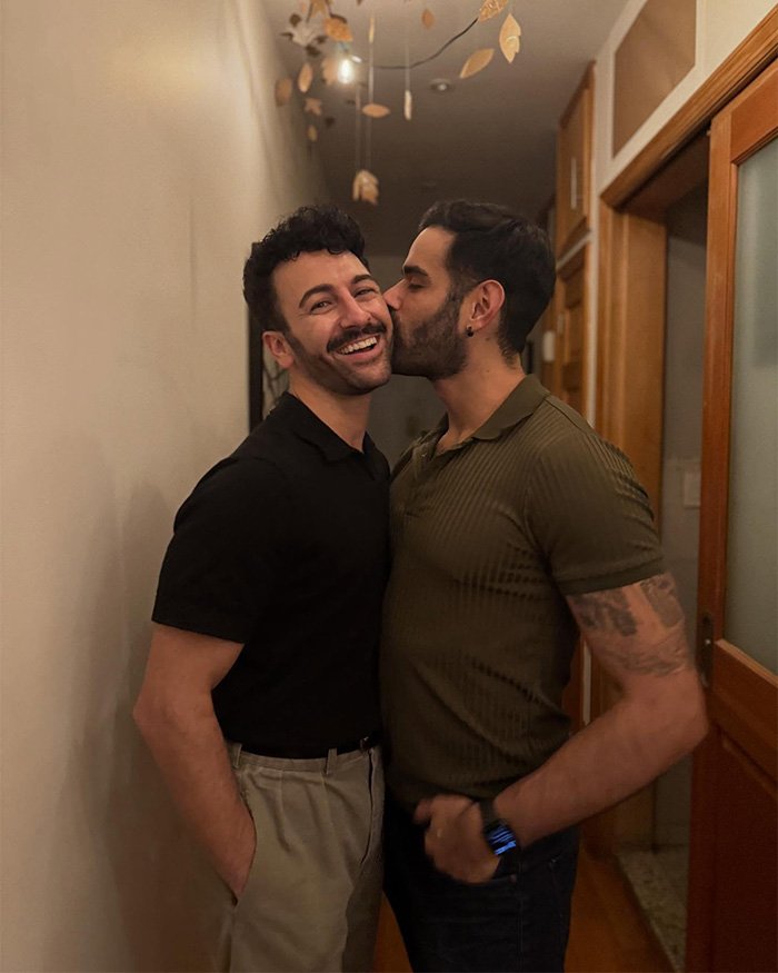 Gay couple in hallway, one kissing partner's cheek as they smile and embrace