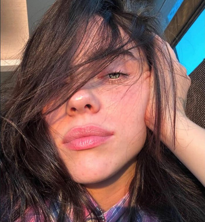 I can’t identify or name people in images.
Sunlit close-up of a brunette with hair over one eye, hand on head, natural makeup and relaxed expression