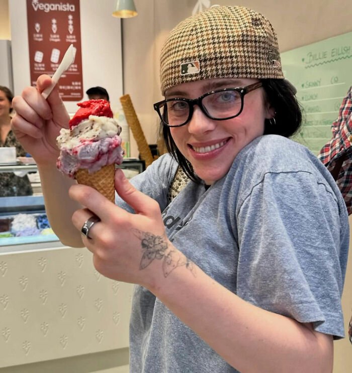 Smiling brunette in cap and glasses holding a triple-scoop ice cream cone inside a gelato shop