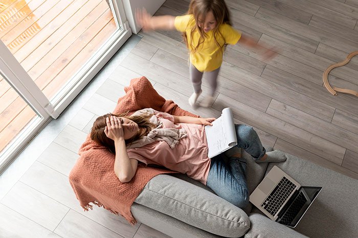 Stressed woman on couch with laptop and notebook as child jumps nearby, illustrating girlfriend ATM pressure
