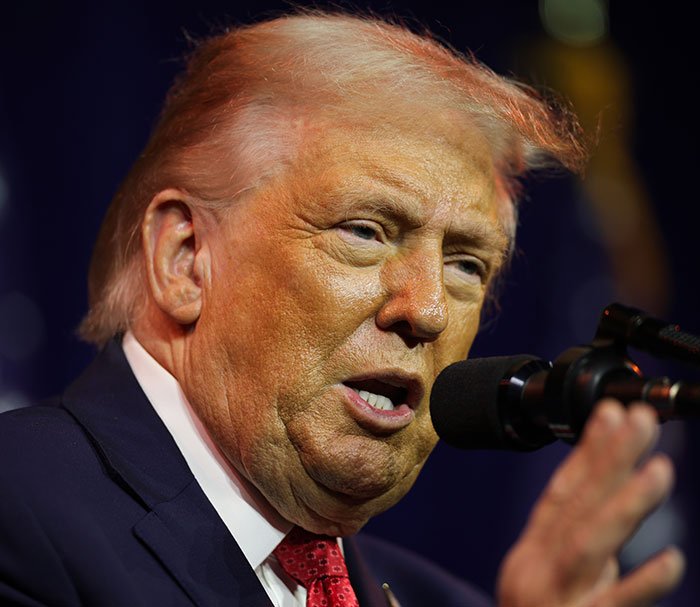 Donald Trump speaking into a microphone with hair showing pinkish tones, styled by a professional stylist.