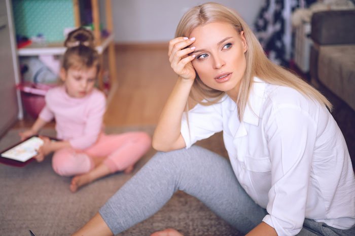 Worried woman sitting on floor while child plays in background, reflecting concern over MIL not changing kid's diaper all day.